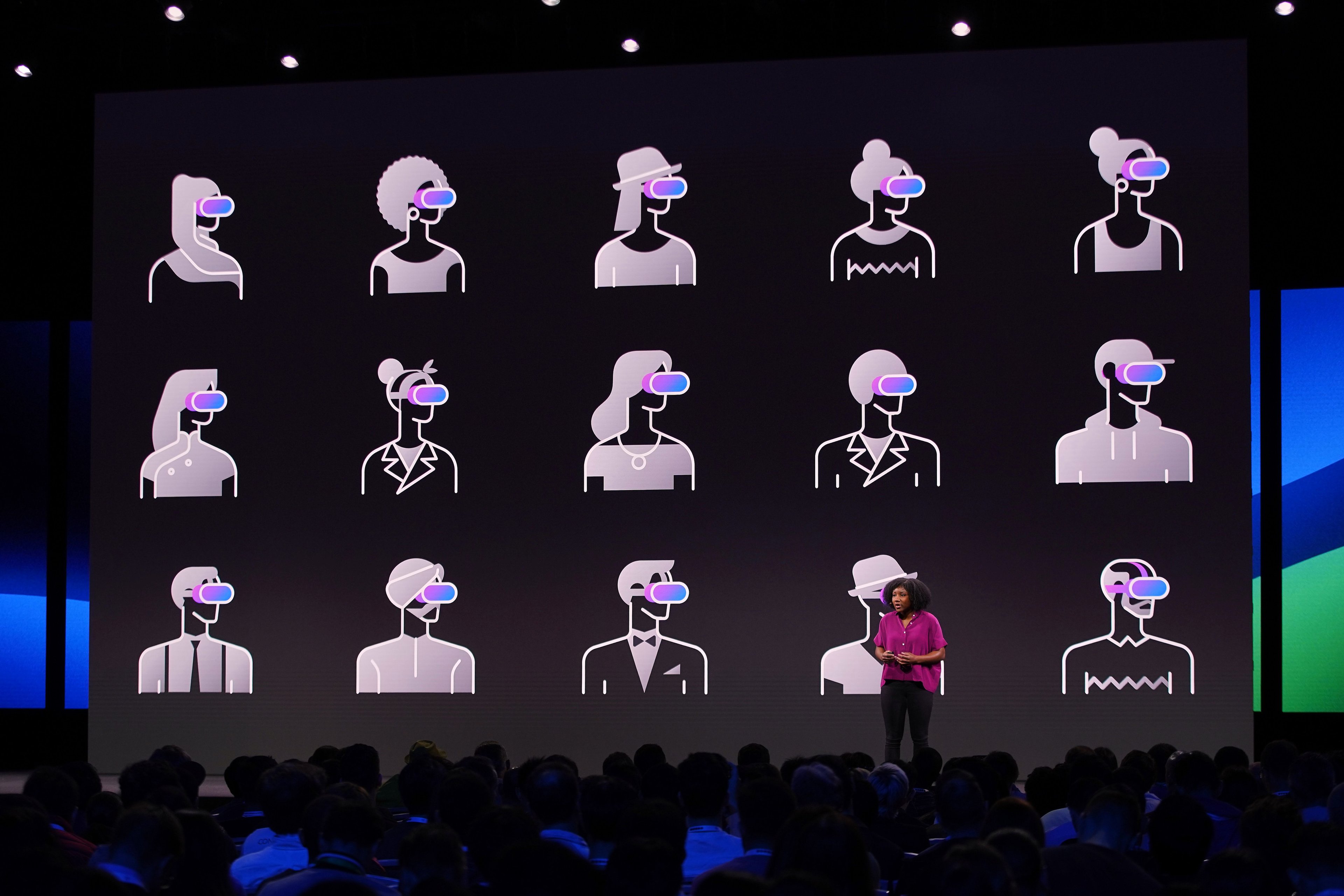A woman presents on stage, with graphics behind her on screen. The graphics are of cartoon humans wearing VR headsets.