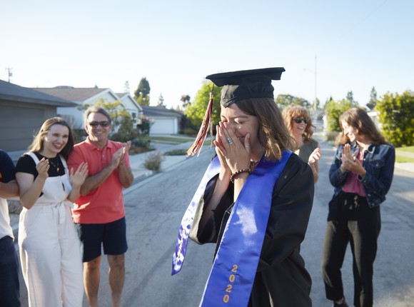 Person in a graduation cap and hat smiling while clapping family and friends surround her. 