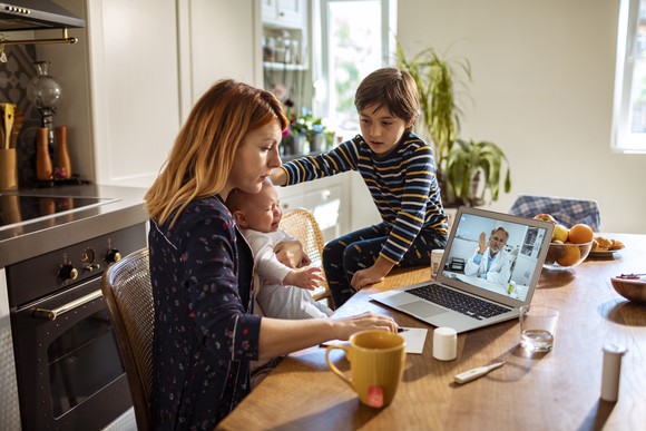 A mom, an infant, and her child sitting at a desk conducting a virtual doctor visit.