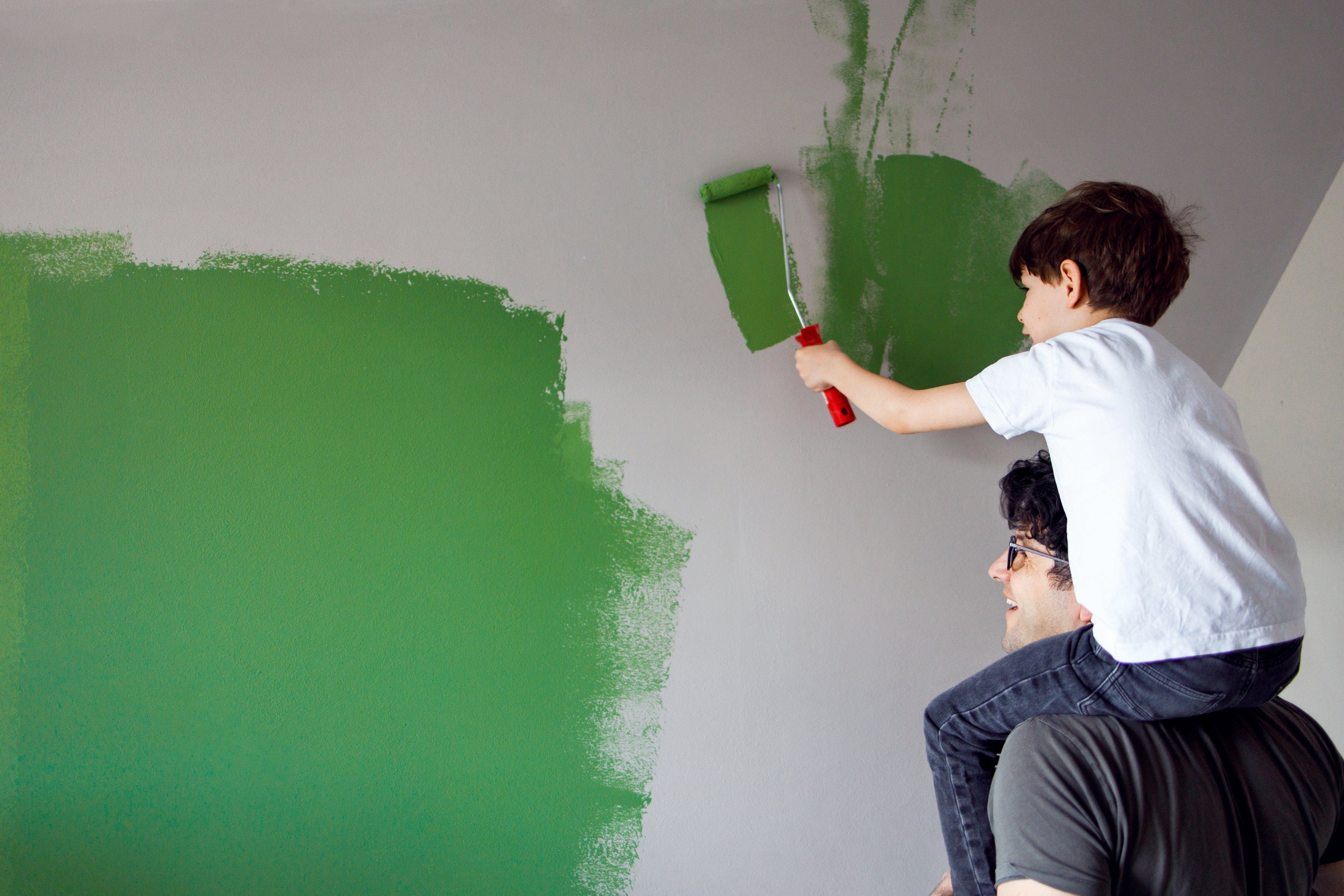 A person holds a child on their shoulders while the child paints a wall green.