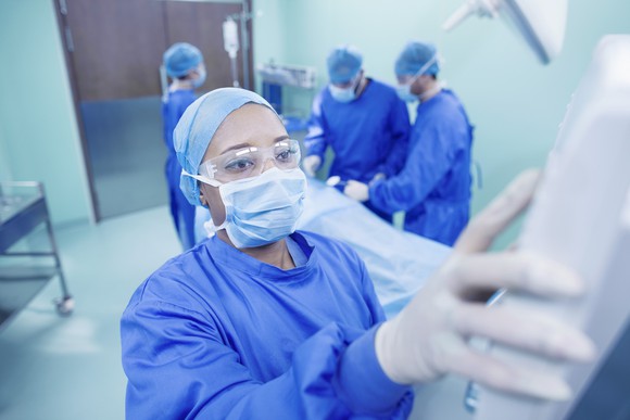 A healthcare worker operates a device inside the operating room with other members of the surgical team in the background.