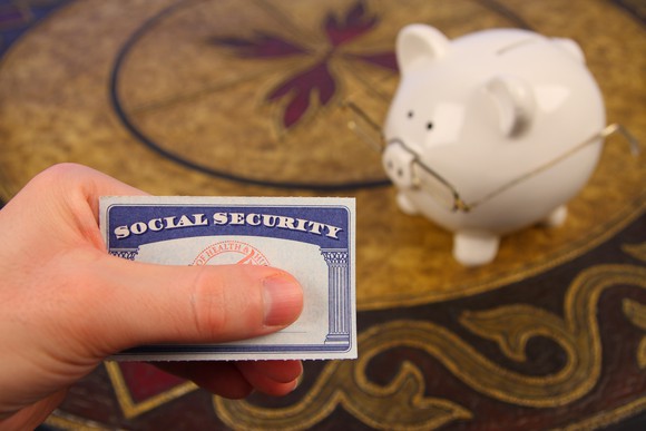 A hand holding a Social Security card next to a white piggy bank wearing glasses.