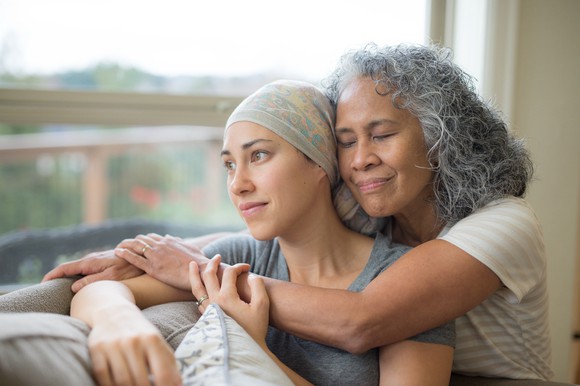 An older person hugging a younger cancer patient from behind as they sit and look out a window.