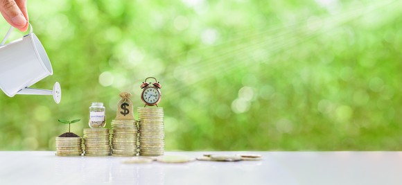 A hand with a watering can is poised near four increasingly larger stacks of coins.