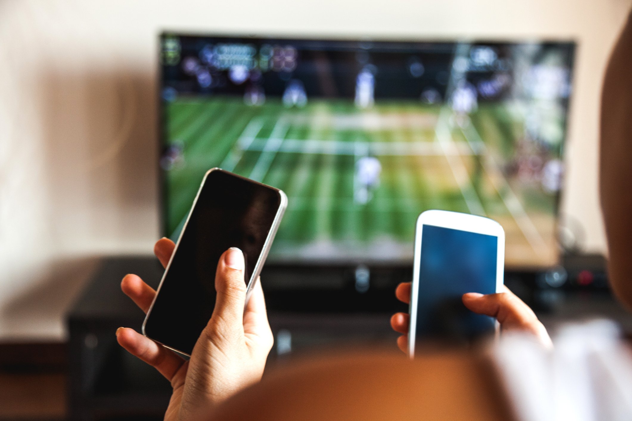 Two people look at cell phones while watching a tennis match on TV. 