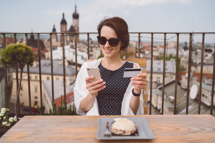 A person seated outdoors in a city coffee shop holding a cell phone and a credit card. 