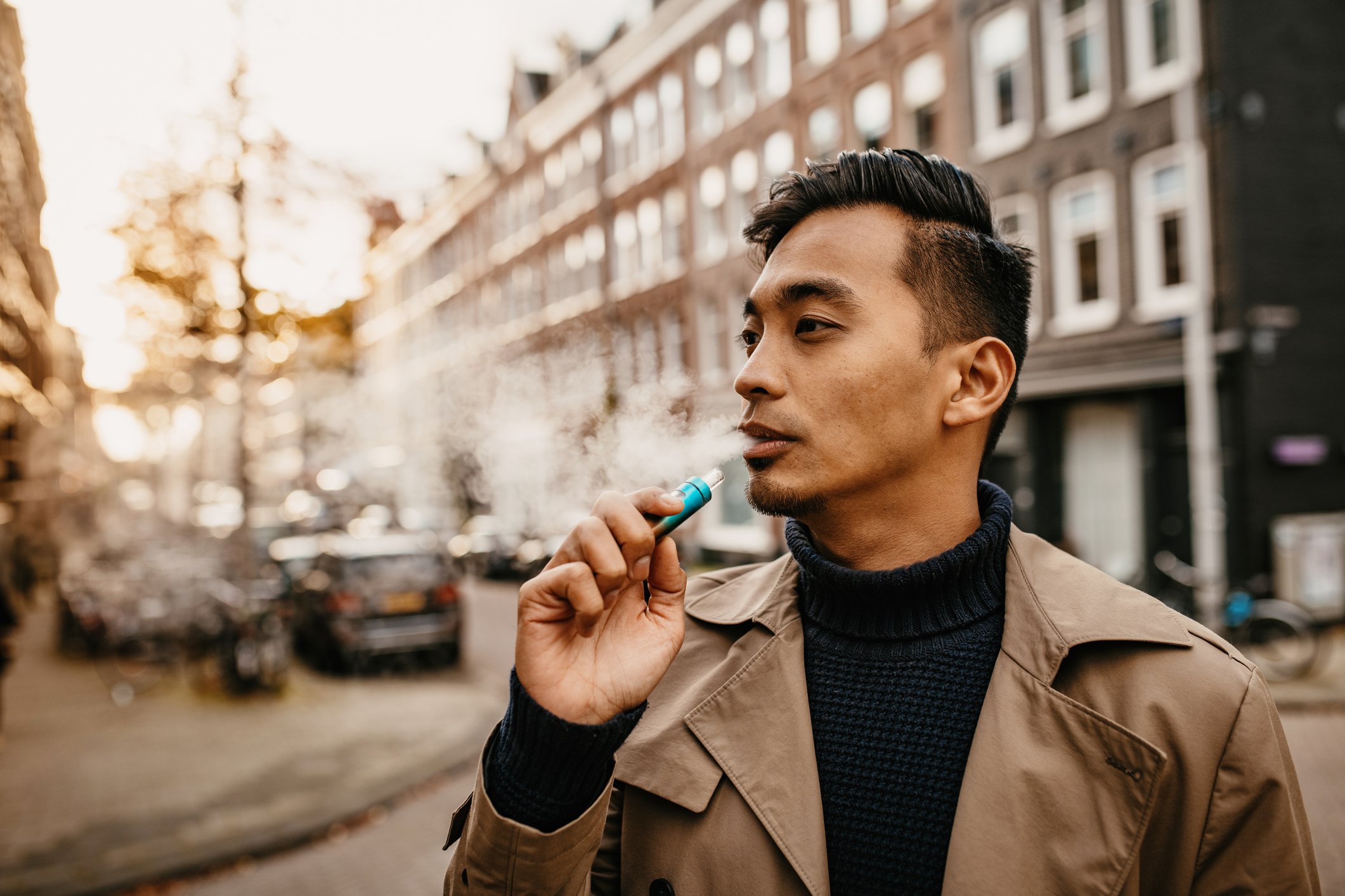 Person looking to the left with a trench coat holding a blue electronic cigarette to their lips.