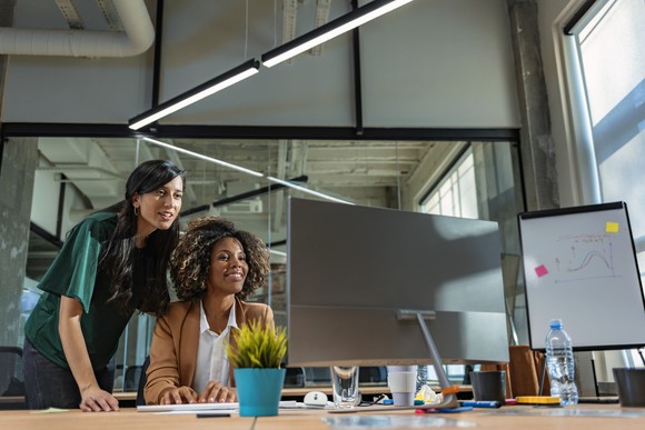 One colleague looks over another's shoulder at a computer screen in an open office with large windows.
