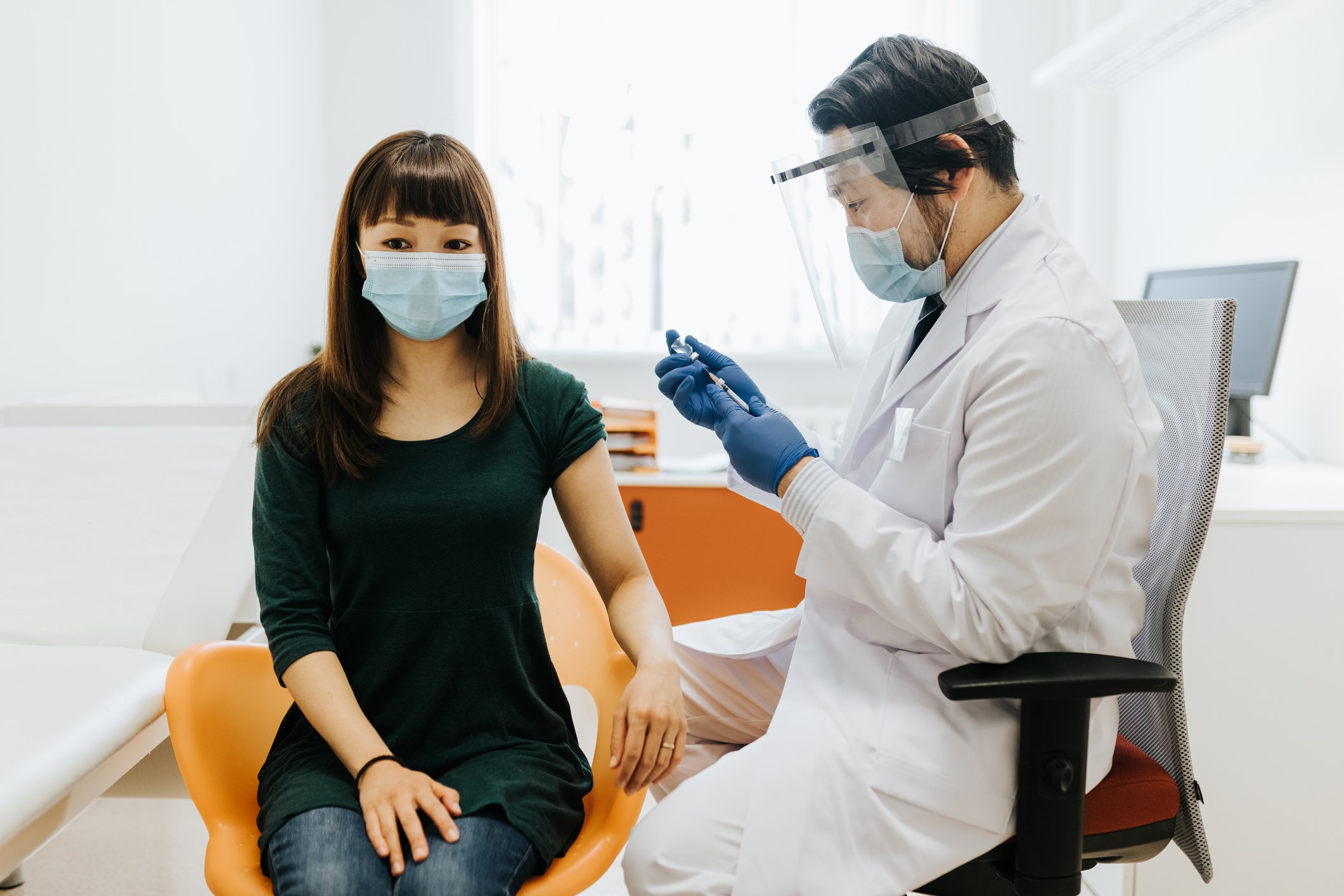 A doctor protected by a face shield prepares a vaccination for a patient.
