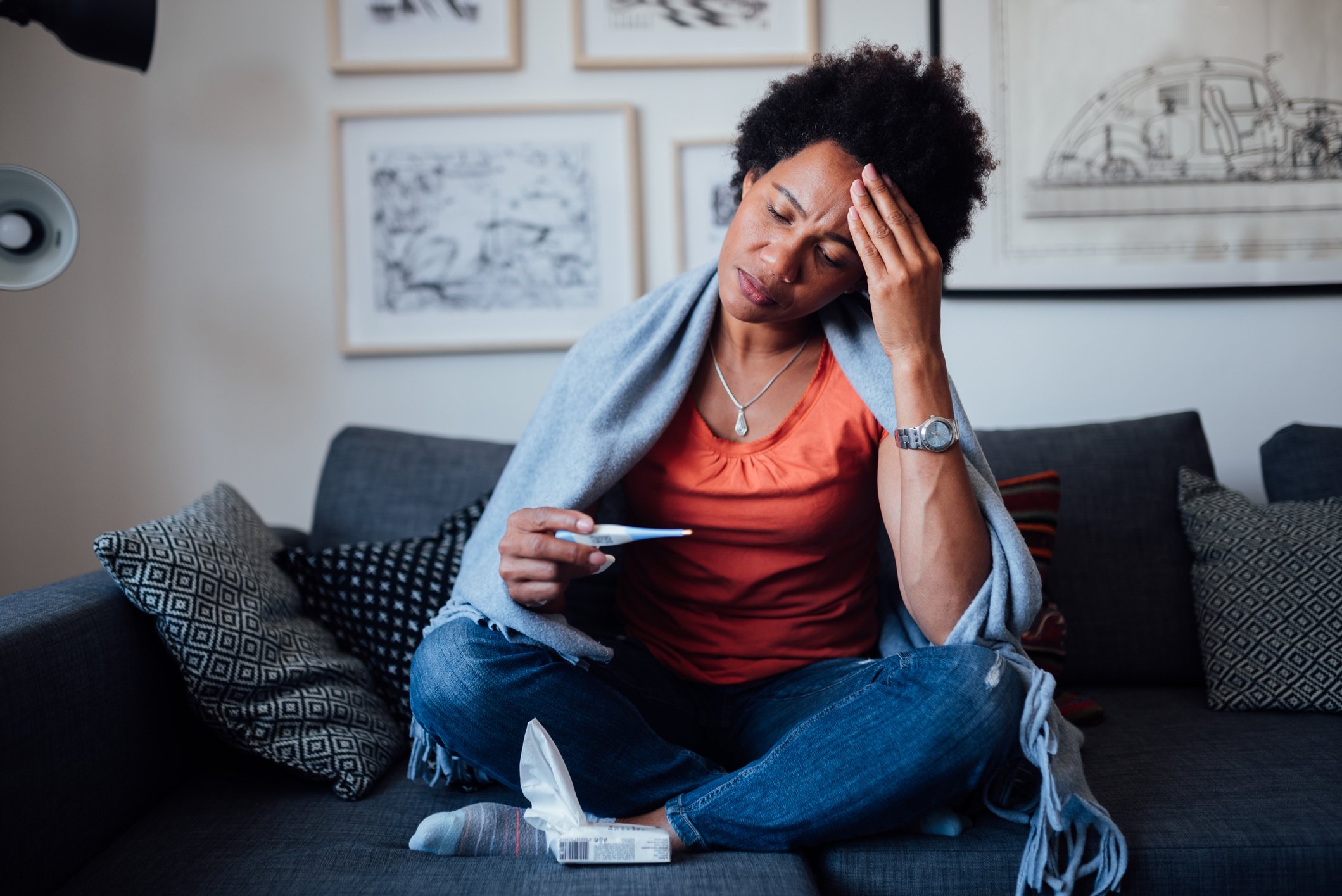 A woman seated on a couch, worrying while looking at a thermometer.
