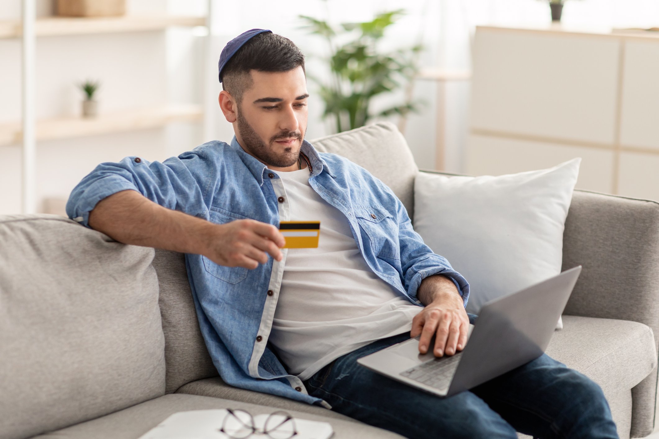 A man sitting on a couch with  computer and holding a credit card.