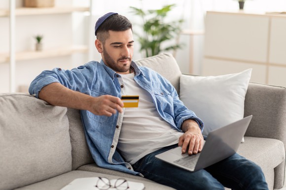 A man sitting on a couch with  computer and holding a credit card.