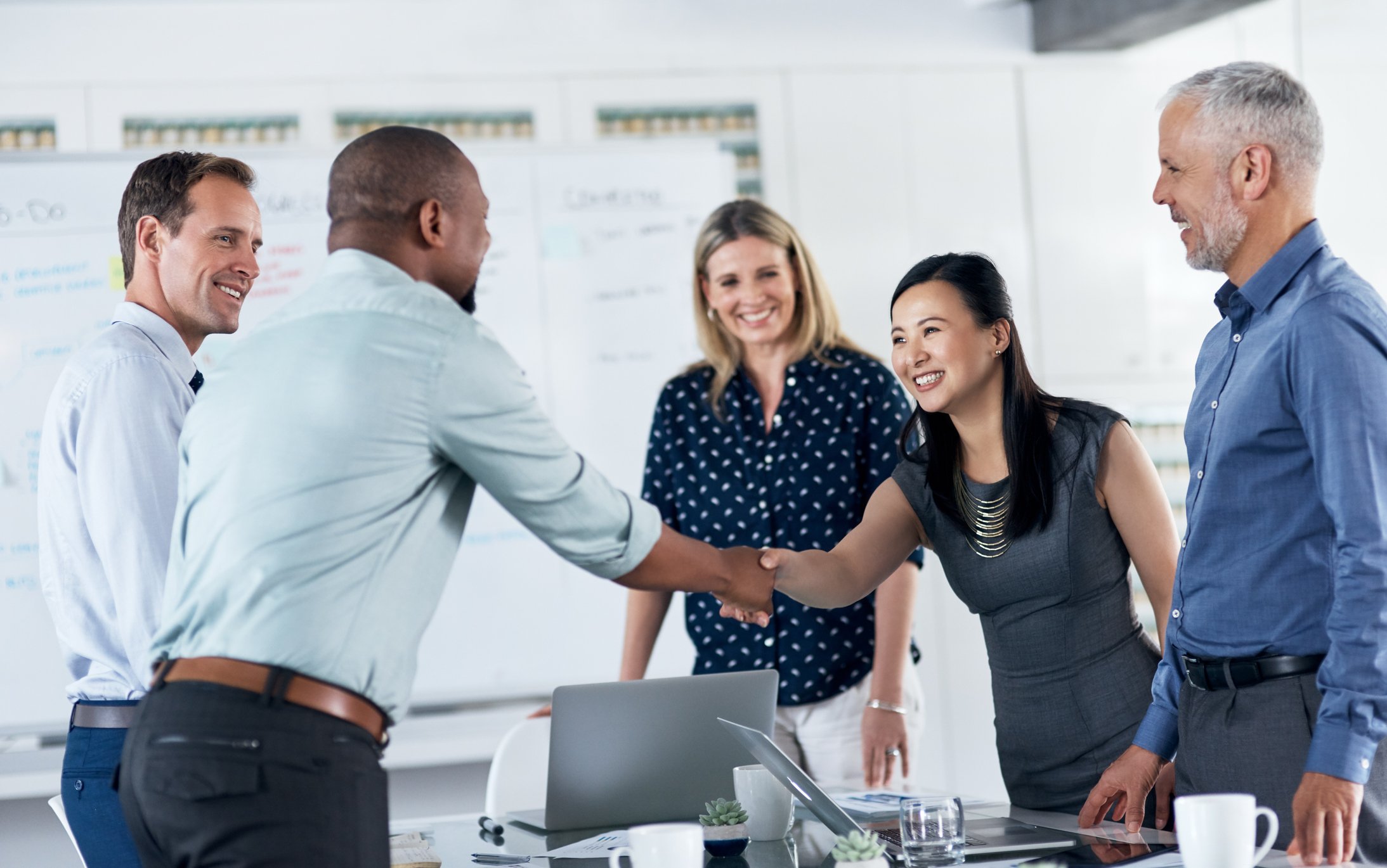 Five people standing around a desk, while two of them shake hands.