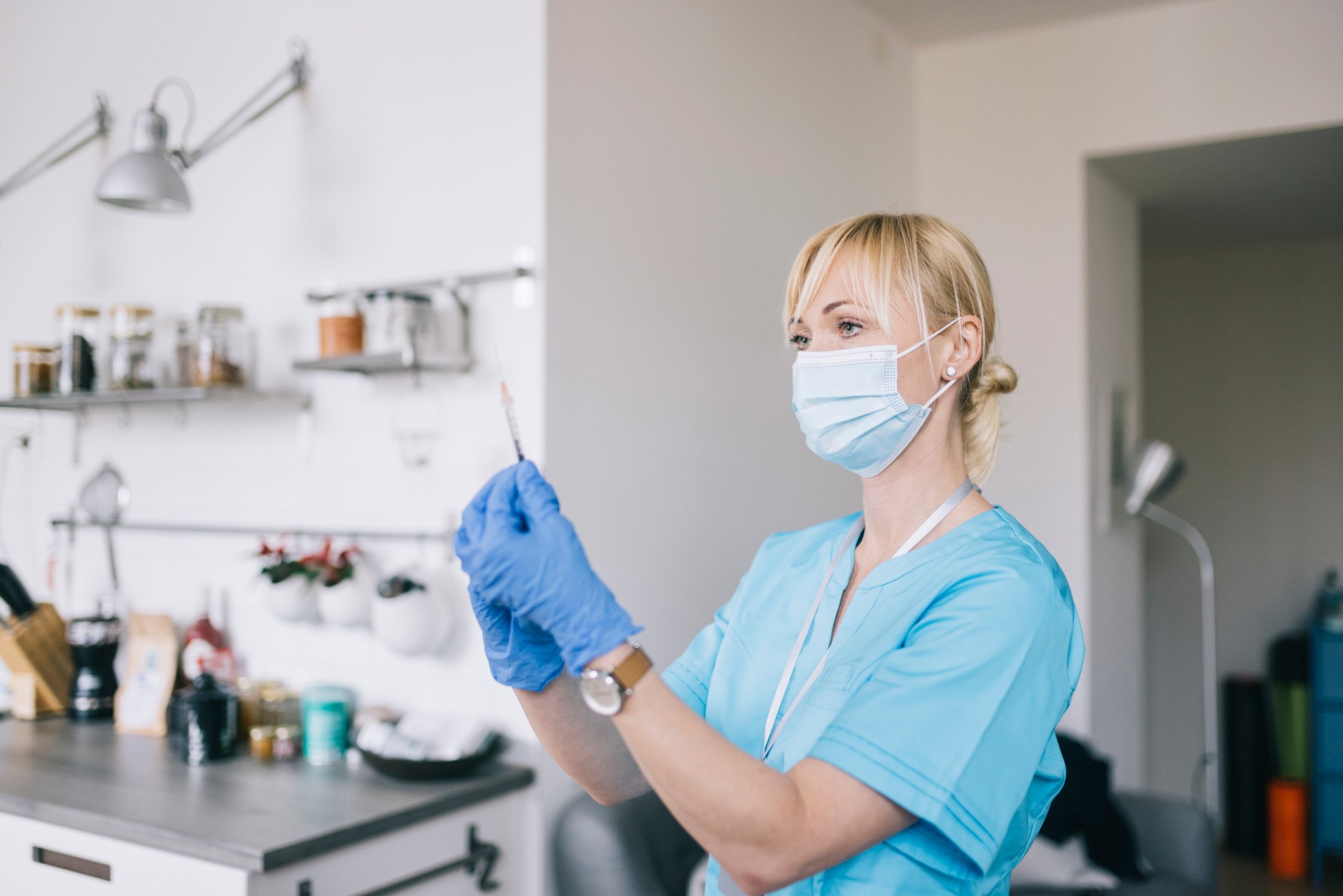 A person wearing scrubs holds a syringe while standing in a kitchen.
