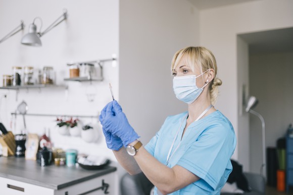A person wearing scrubs holds a syringe while standing in a kitchen.