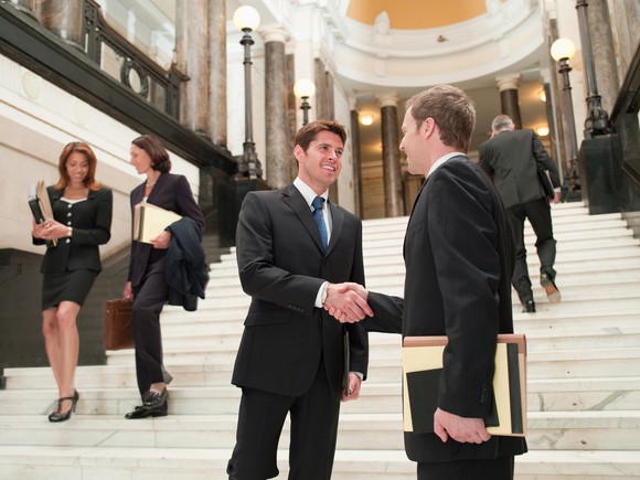 Two businesspeople shaking hands on the steps inside the entrance of a large building.
