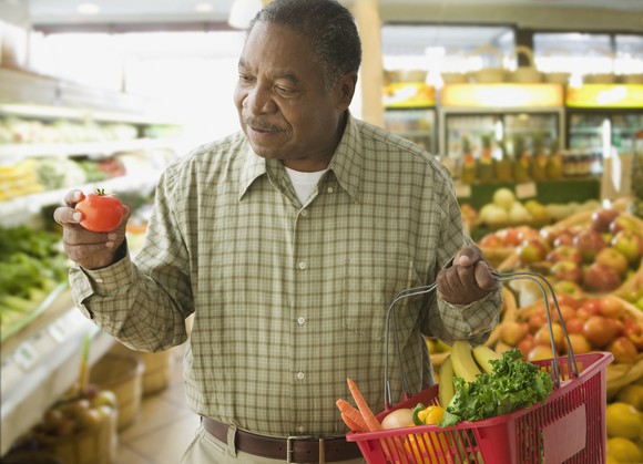Someone in a grocery store looking at the tomato in his hand while holding a basket of produce in his other hand. 