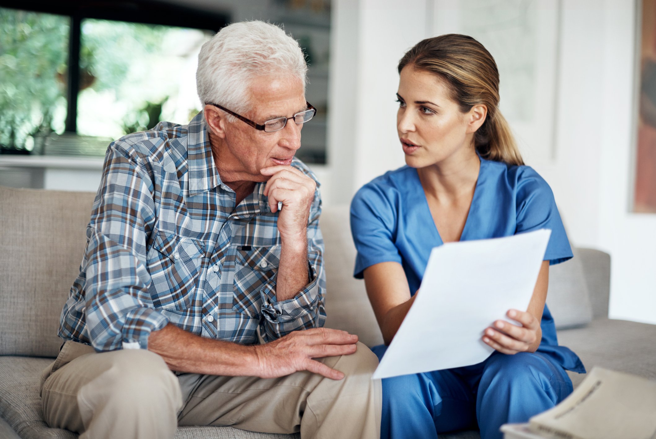 A doctor and a patient sit on a couch together as the doctor holds up a piece of paper and explains something to the patient.