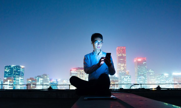 A young man operates his cell phone sitting on a rooftop at night in front of a skyline. 