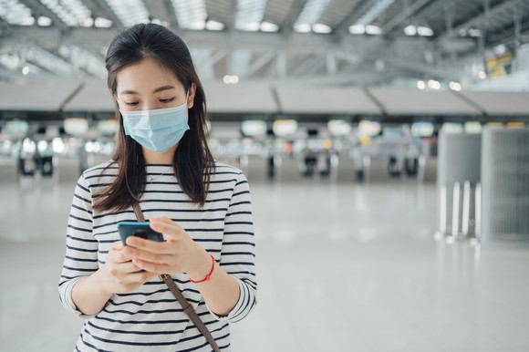 Young woman in face mask operates her phone in an airport. 