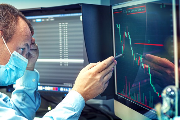 A worried business person in a protective mask studies a falling stock price chart on a computer screen.