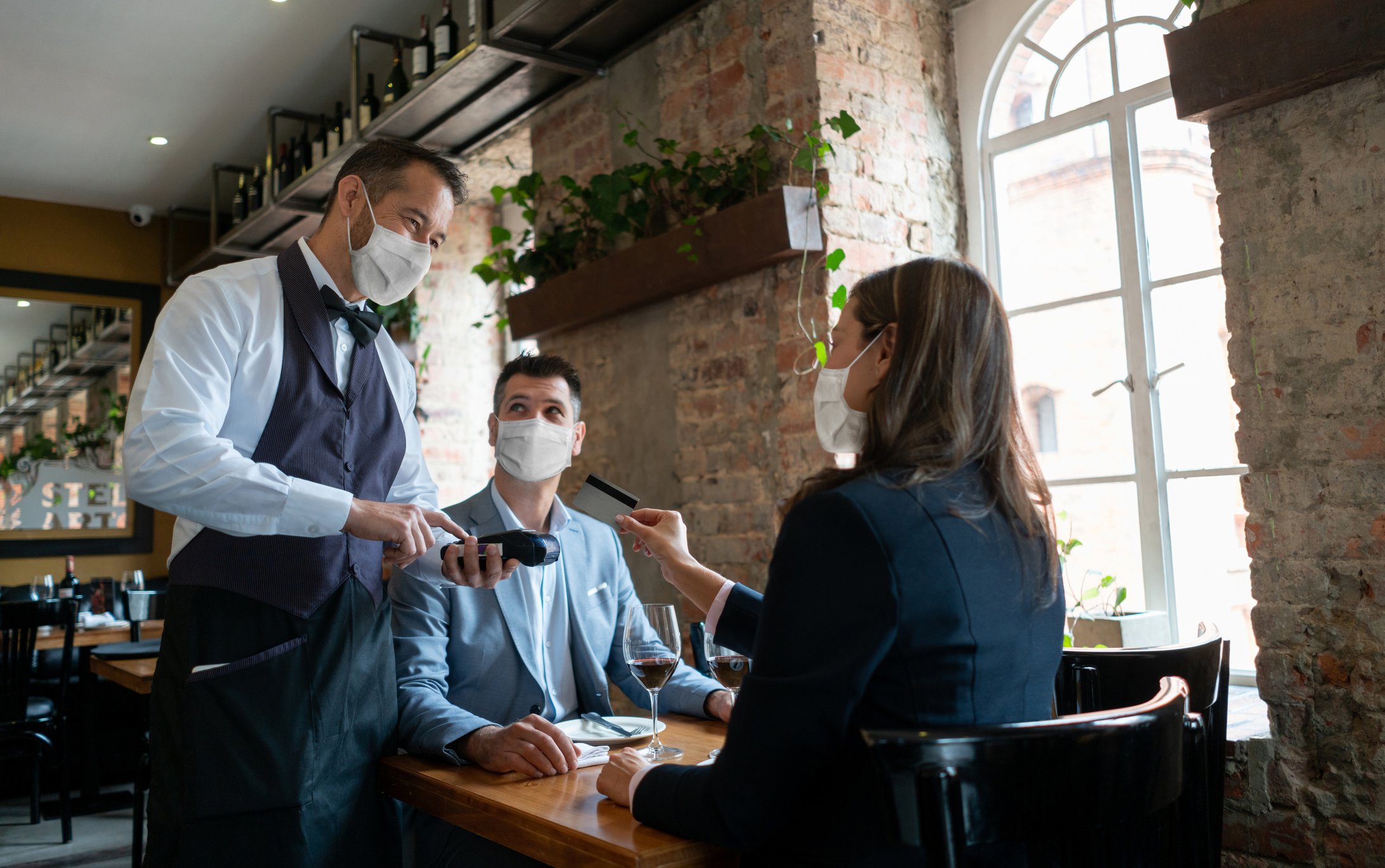 A waiter taking a credit card from two patrons sitting at a table inside a restaurant.