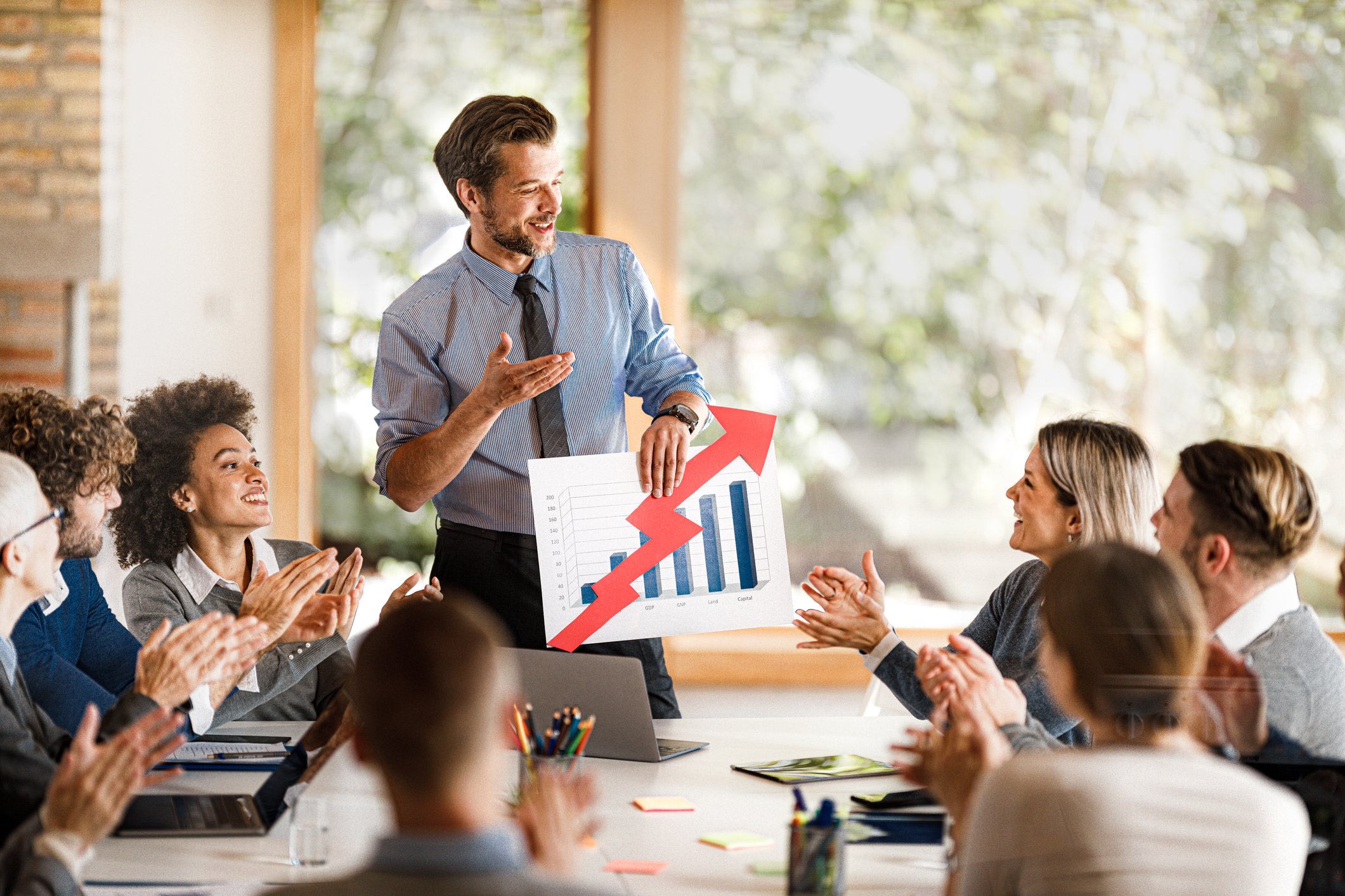 Person holding chart showing soaring numbers, with others seated at a table.