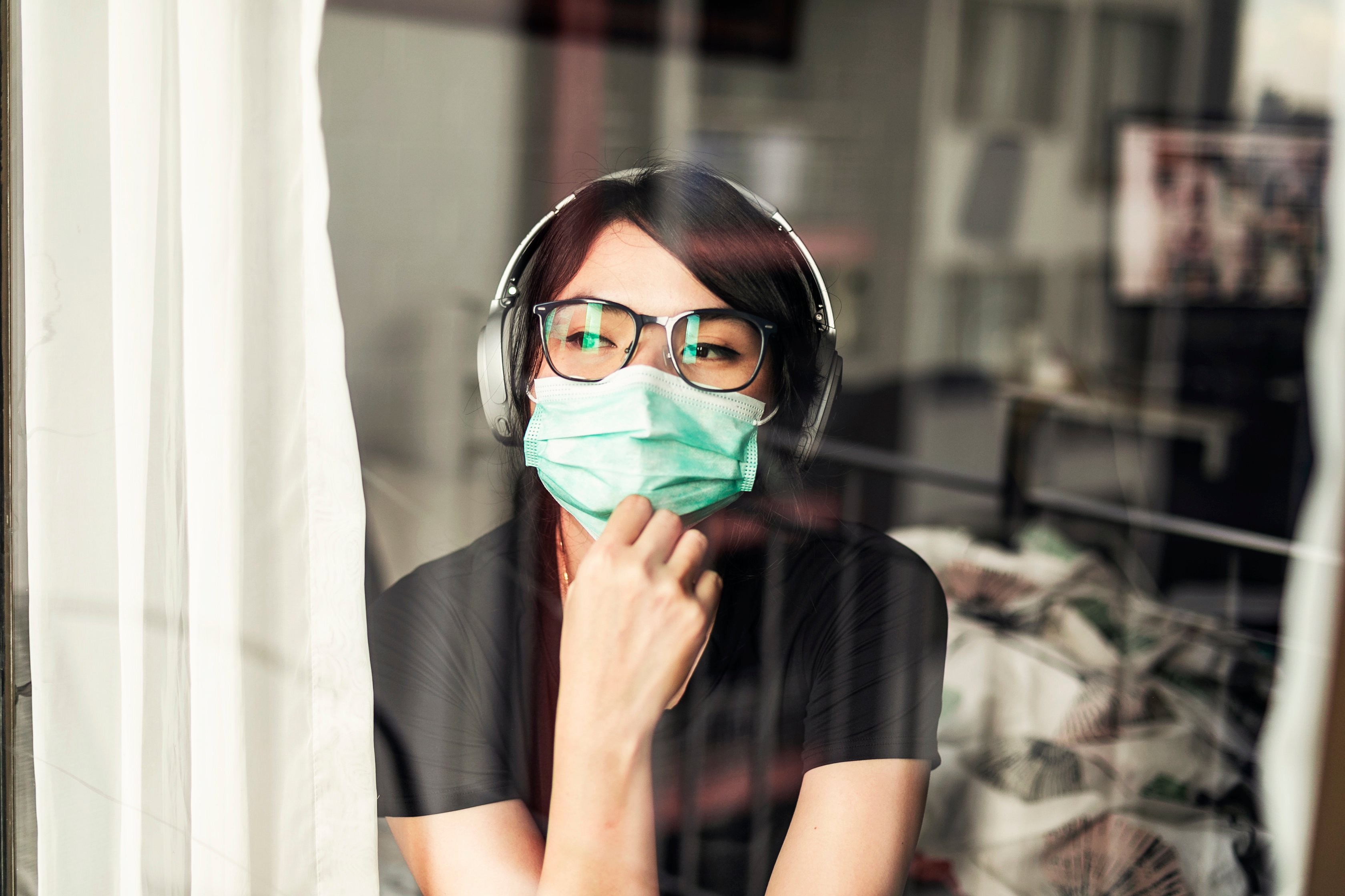 A person wearing a surgical mask and headphones looks out at the world through a curtained window.