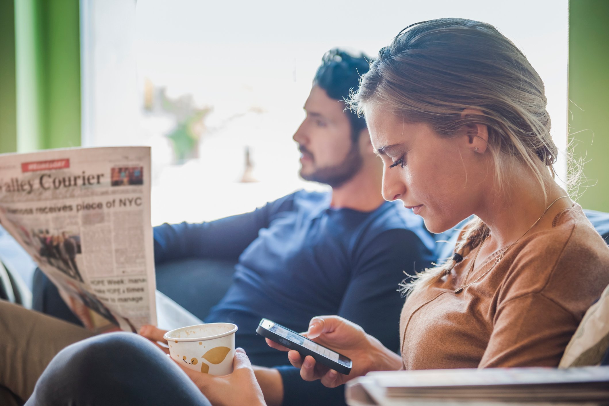 Two people, one of which is reading a newspaper, the other looking at a smartphone.