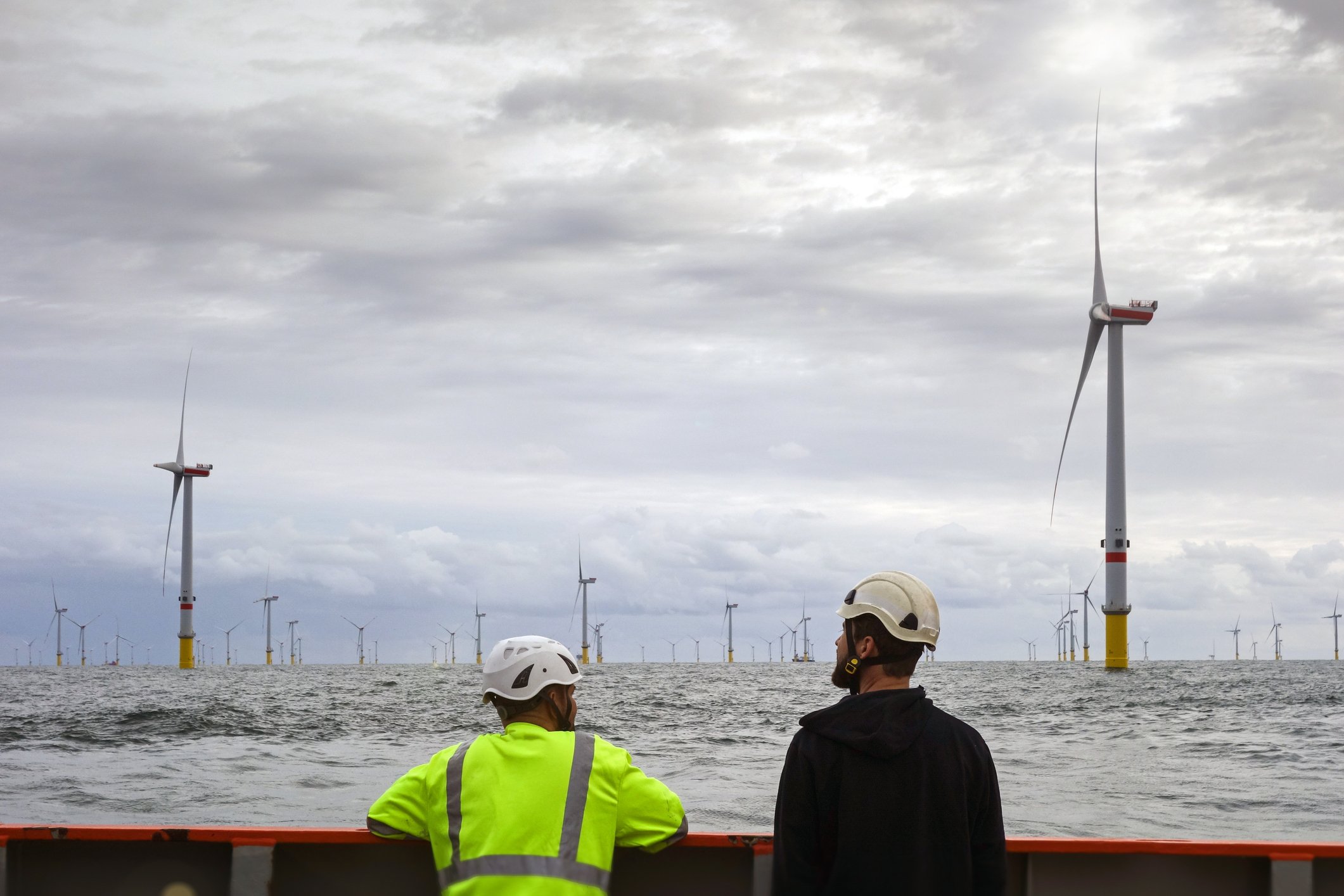 Two workers in hard hats look at an offshore wind energy farm from a vessel. 