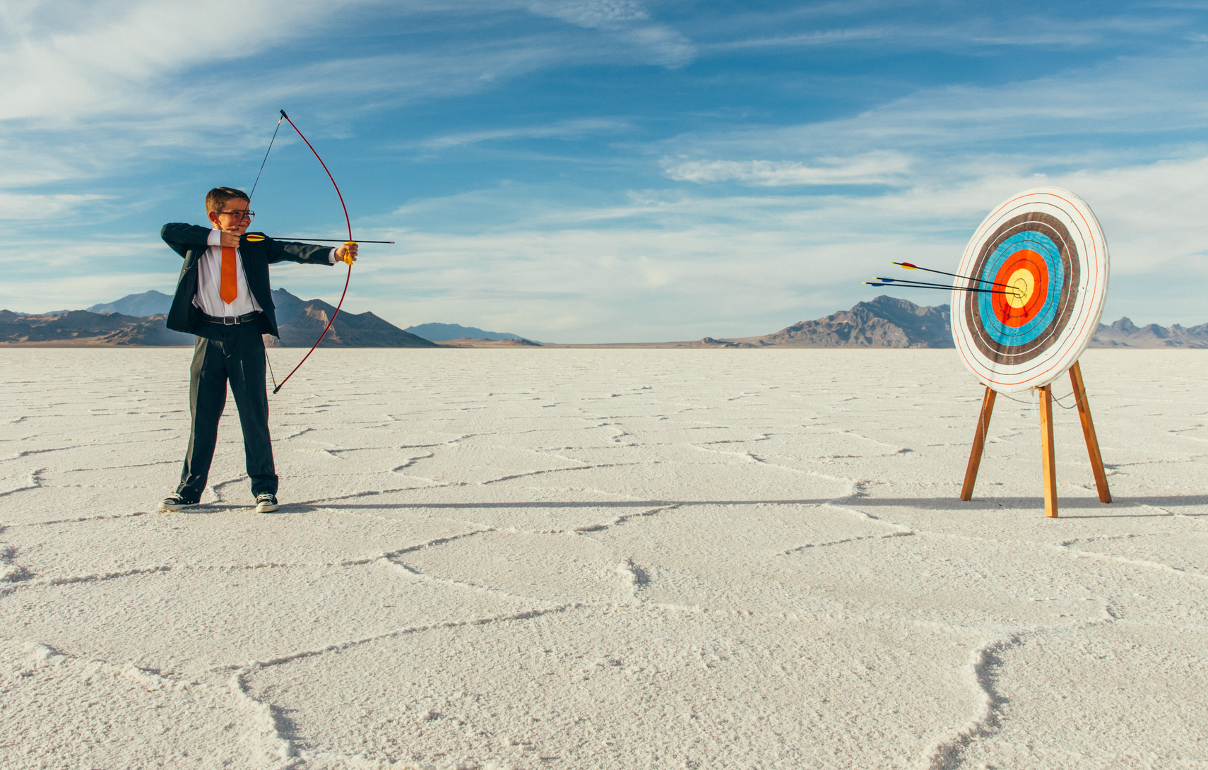 a young man in a business suit prepares to shoot an arrow from a bow at a nearby target while standing on white sands in a desert setting with mountains and blue skies in the background
