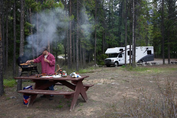A camper cooking food on a campground table with a white RV parked among pines in the background.