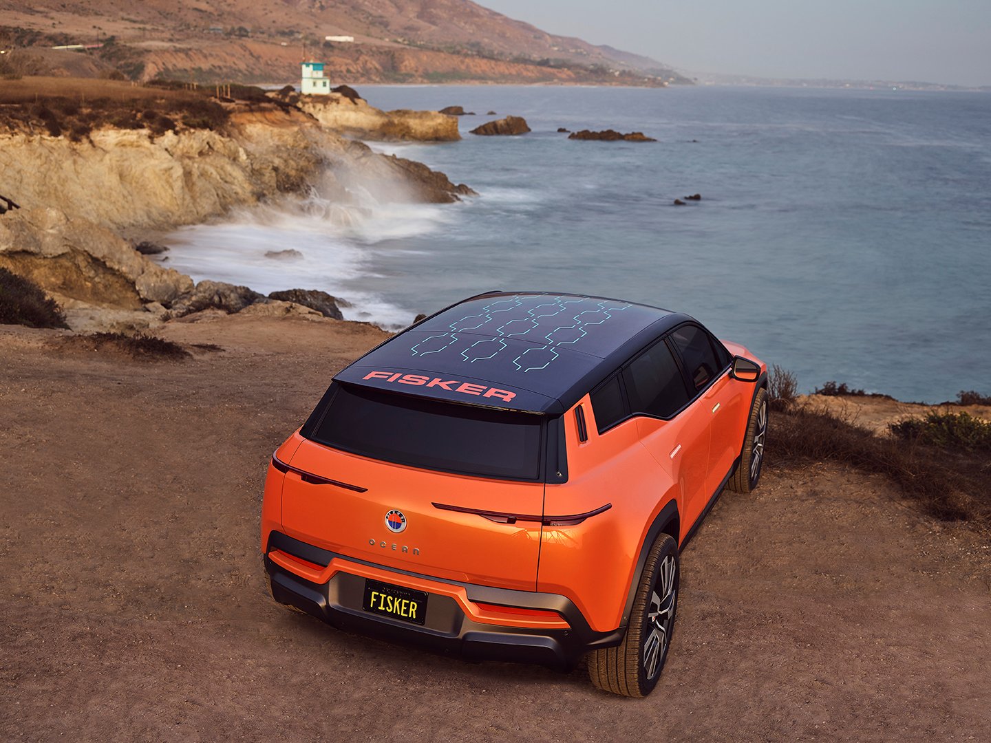 Orange Fisker Ocean SUV on a cliff facing the ocean. 