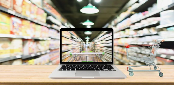 A view of a grocery store aisle through a transparent laptop screen beside a miniature shopping cart.