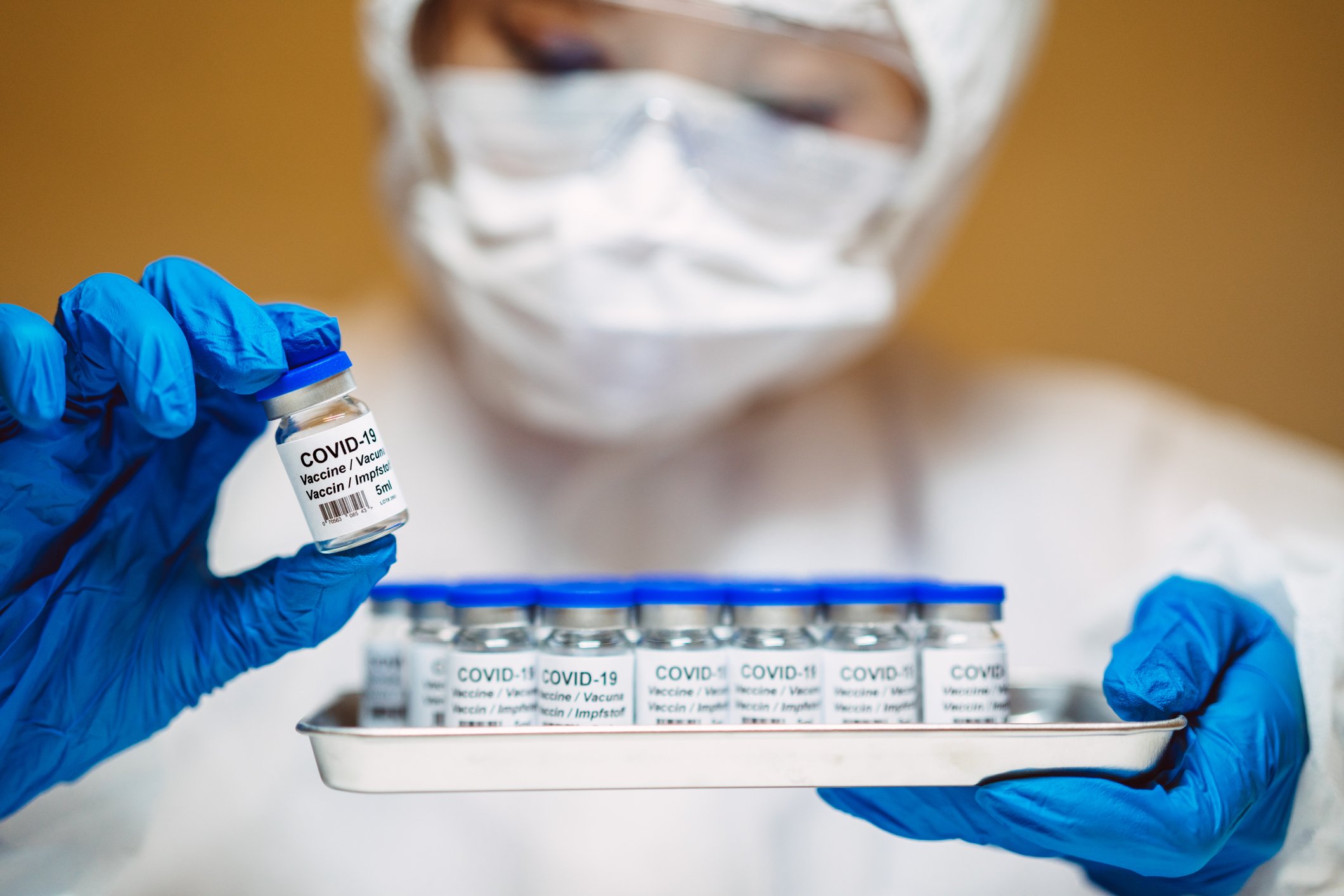 A person in full personal protective equipment, holding a rack of glass vials labeled COVID-19 Vaccine in multiple languages.