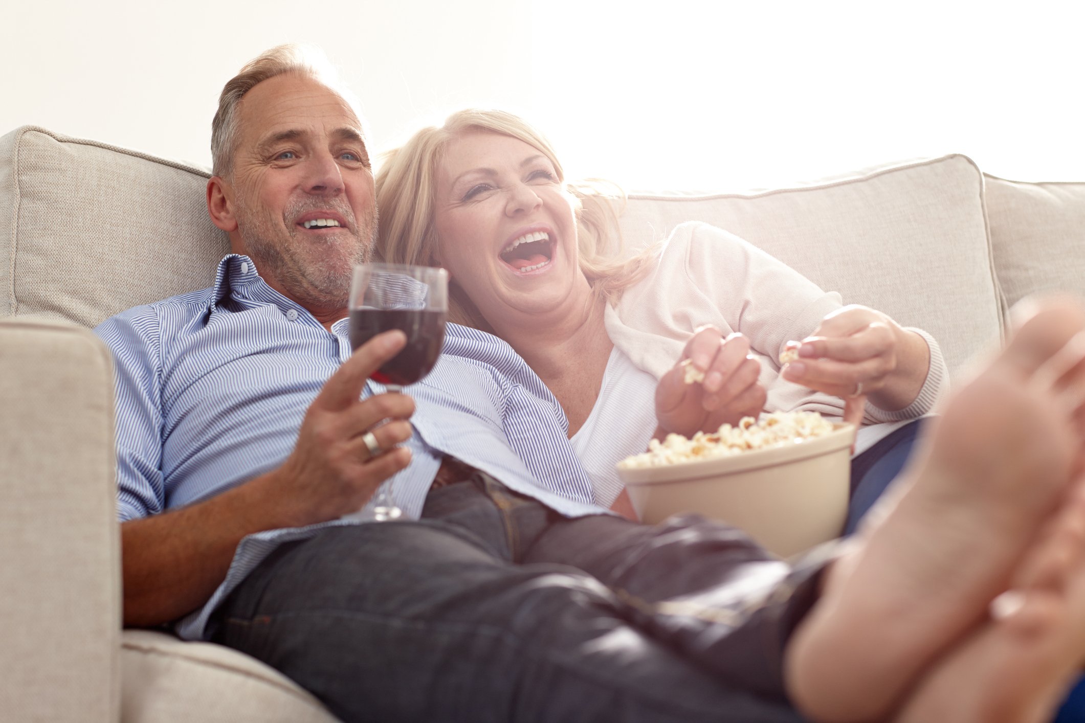 Two smiling people share popcorn and wine, snuggled up on a couch.