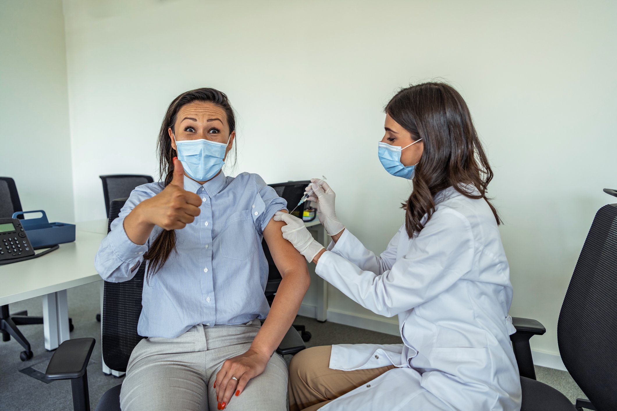 A patient gives an enthusiastic thumbs-up and smiles behind a mask while receiving a shot from a masked healthcare worker.