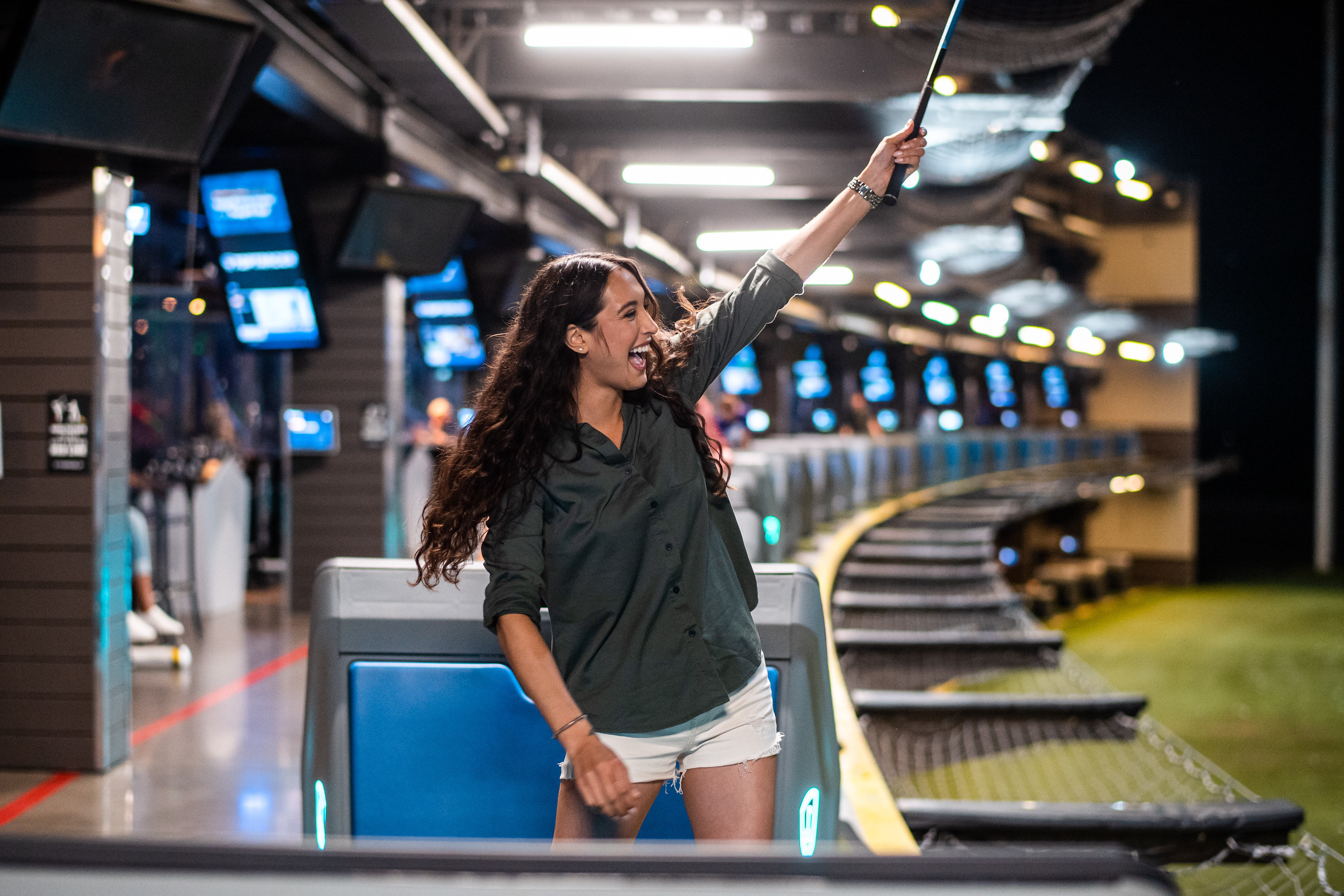 A woman looks out on the field of a Top Golf facility in Texas while standing in a golf box and holding a golf club