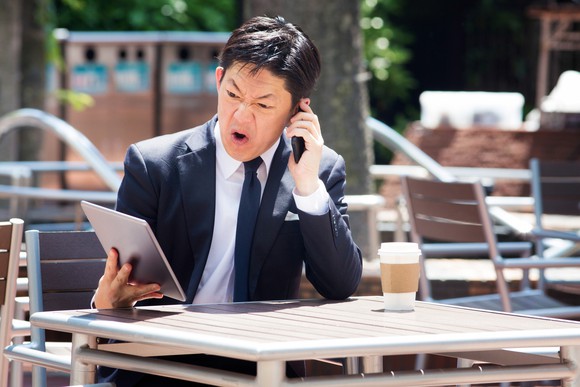 A person sitting at an outdoor table with coffee, looking at a tablet in disgust while talking on the phone