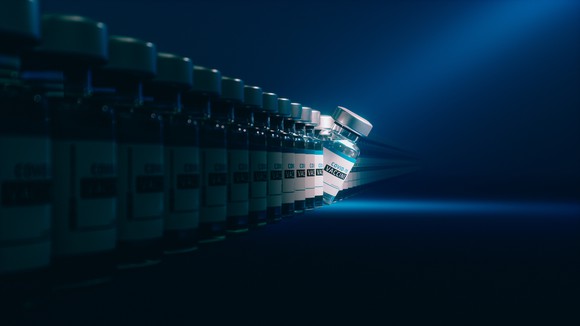 A line of vaccine vials labeled COVID-19 Vaccine, with one tipping over and a light shining on it.