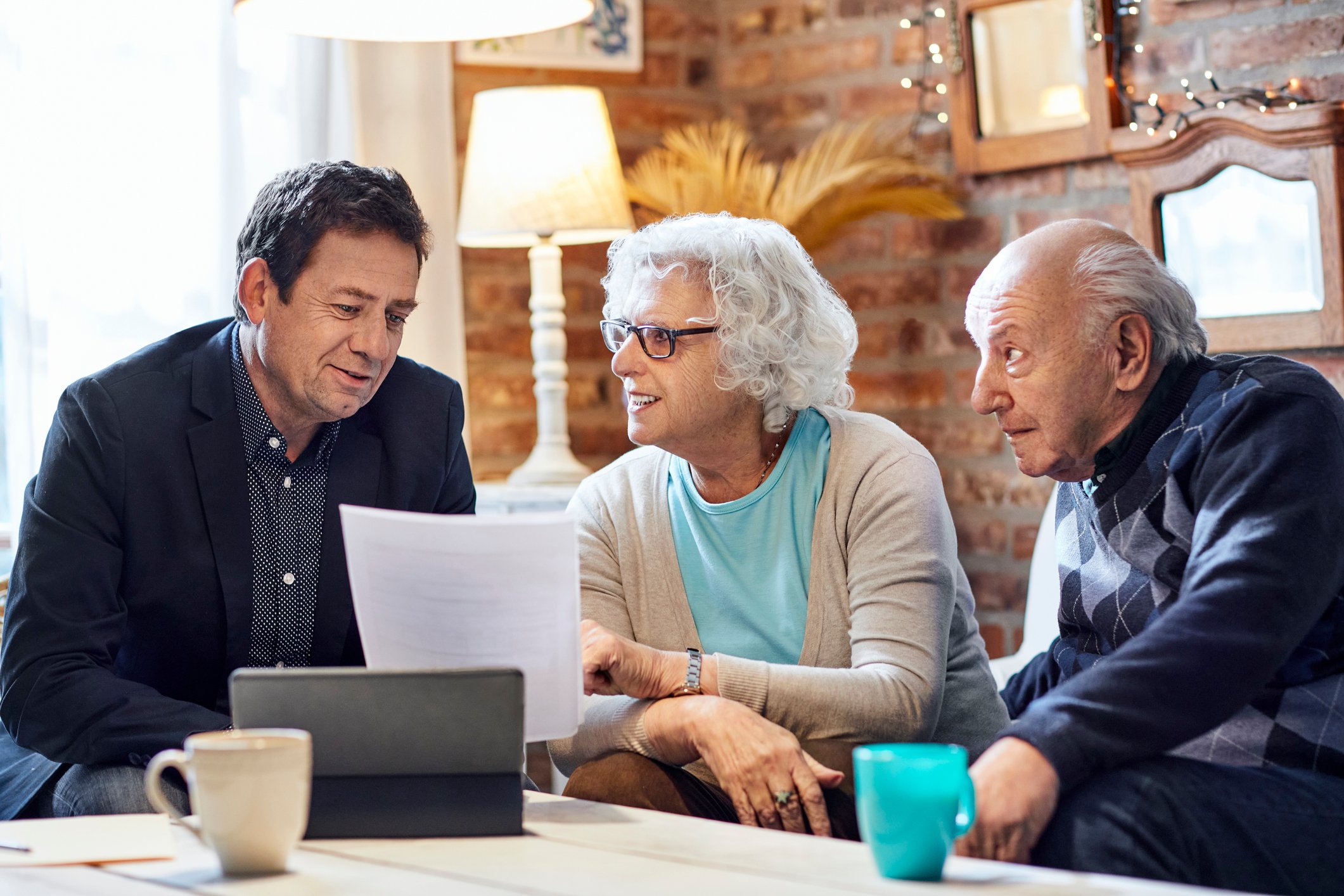 An older couple talks to a man in a suit jacket while all three sit at a coffee table and look at documents.