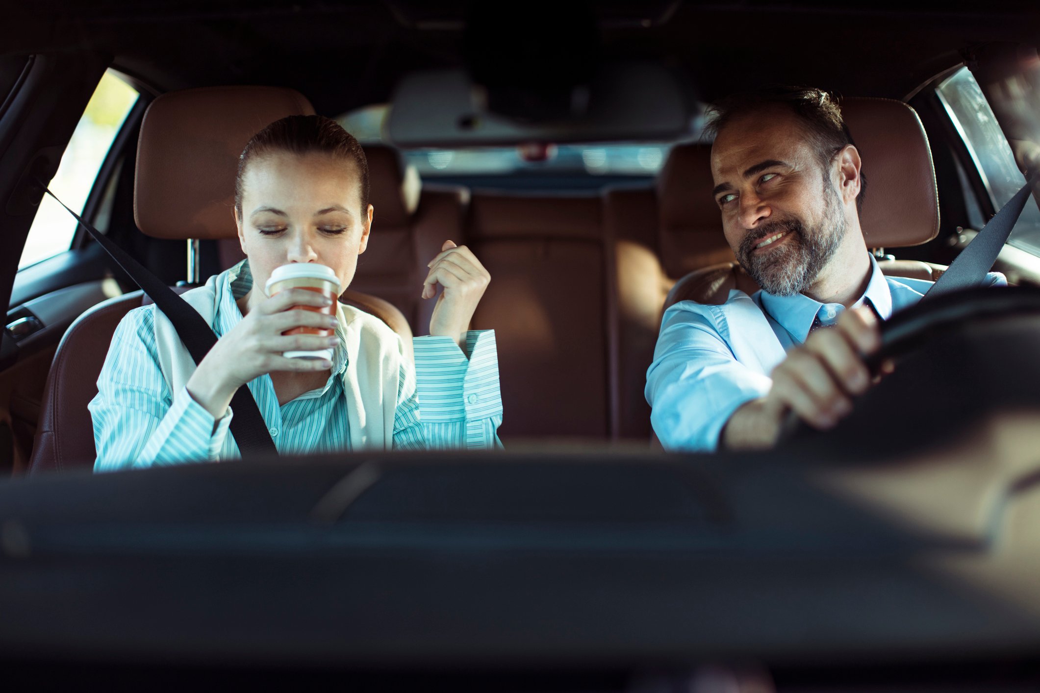 Two people sitting in a car and the person in the passenger's seat is drinking a beverage.