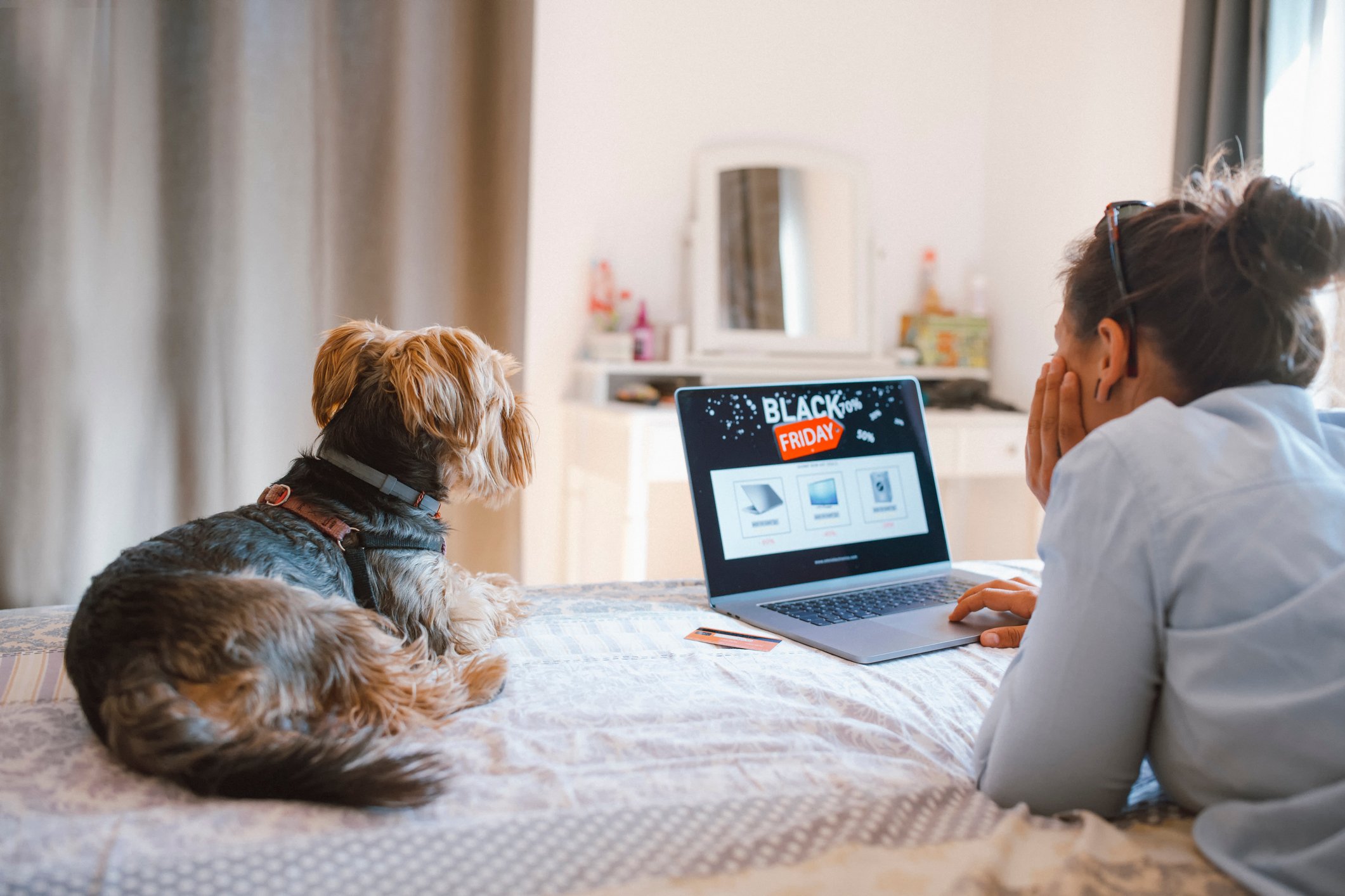 A person lies on their bed with a dog looking at a website listing a Black Friday sale.