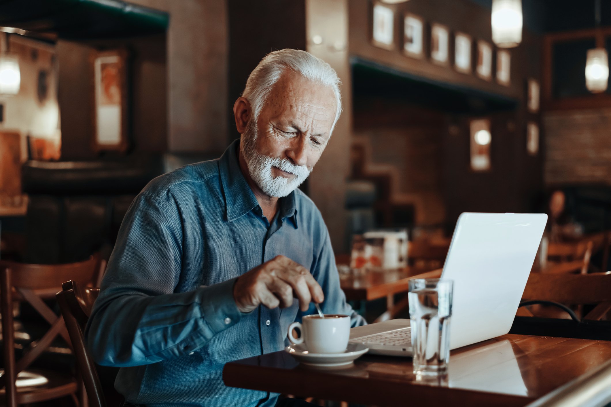 Senior sitting in coffee shop with laptop and cup of coffee.
