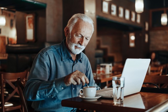 Senior sitting in coffee shop with laptop and cup of coffee.
