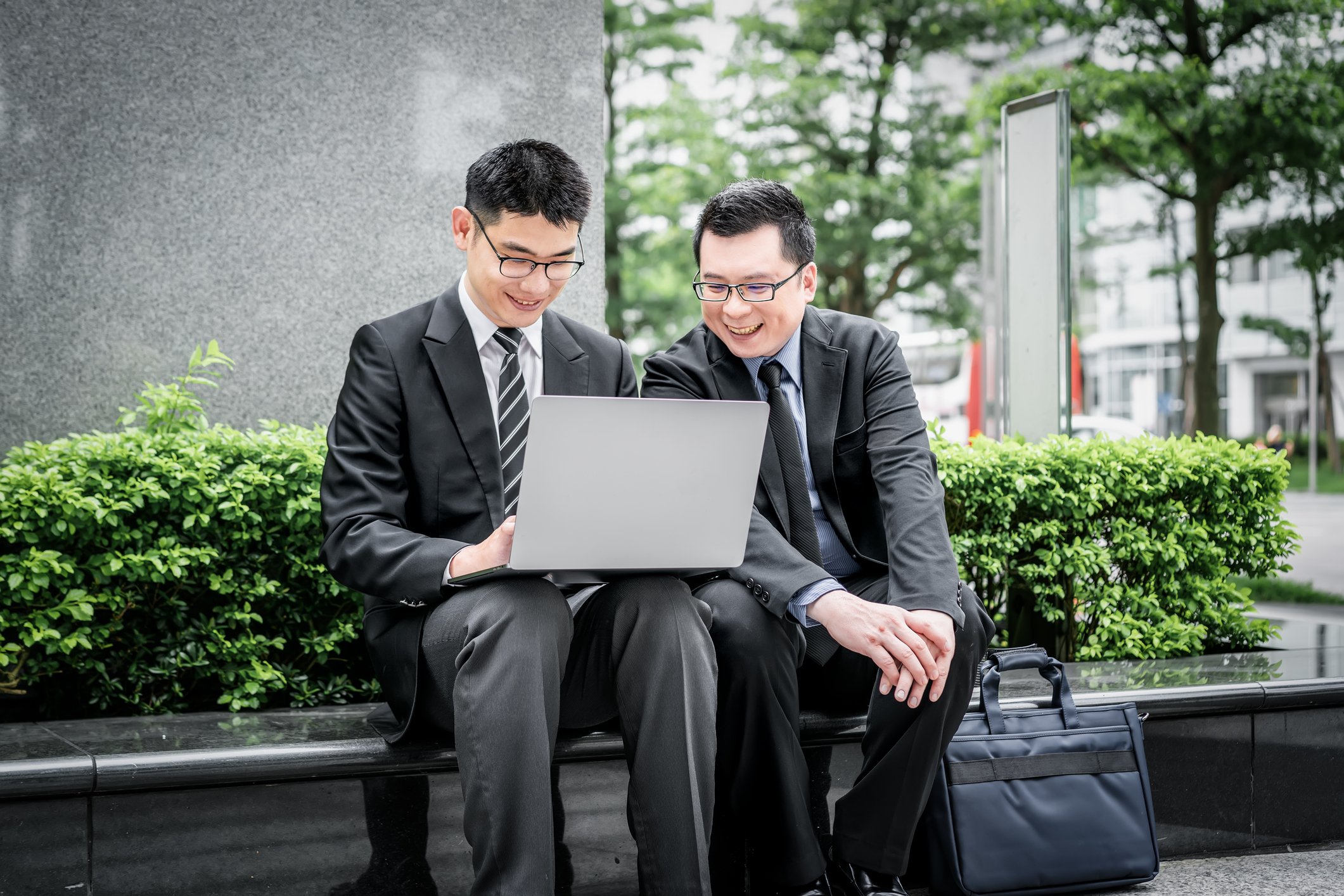 Two businessmen sit on a bench looking at a laptop, smiling. 