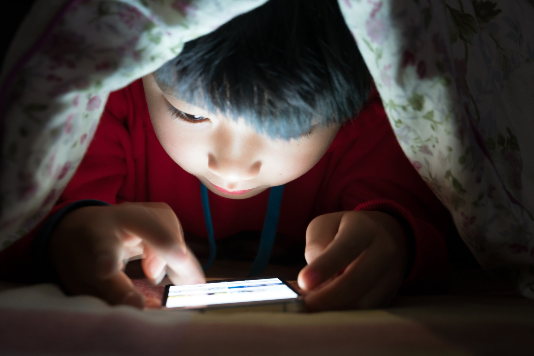 A young boy looks at a smartphone under his covers. 