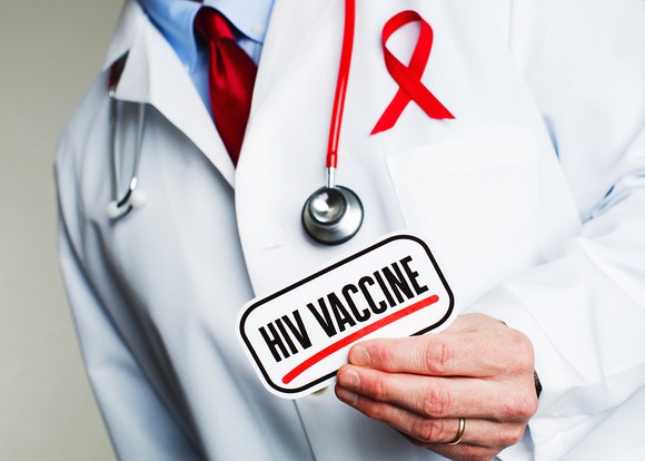 A person wearing a white lab coat with a red ribbon in the pocket holding a card with "HIV vaccine" printed on it.