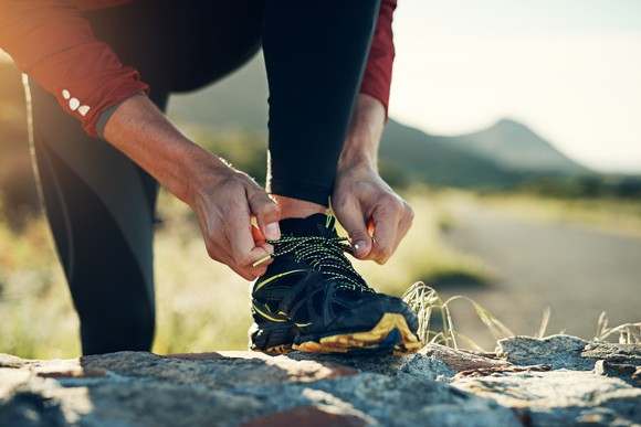 A runner outdoors, ensuring their athletic shoes' laces are tied, with their foot propped on a boulder.