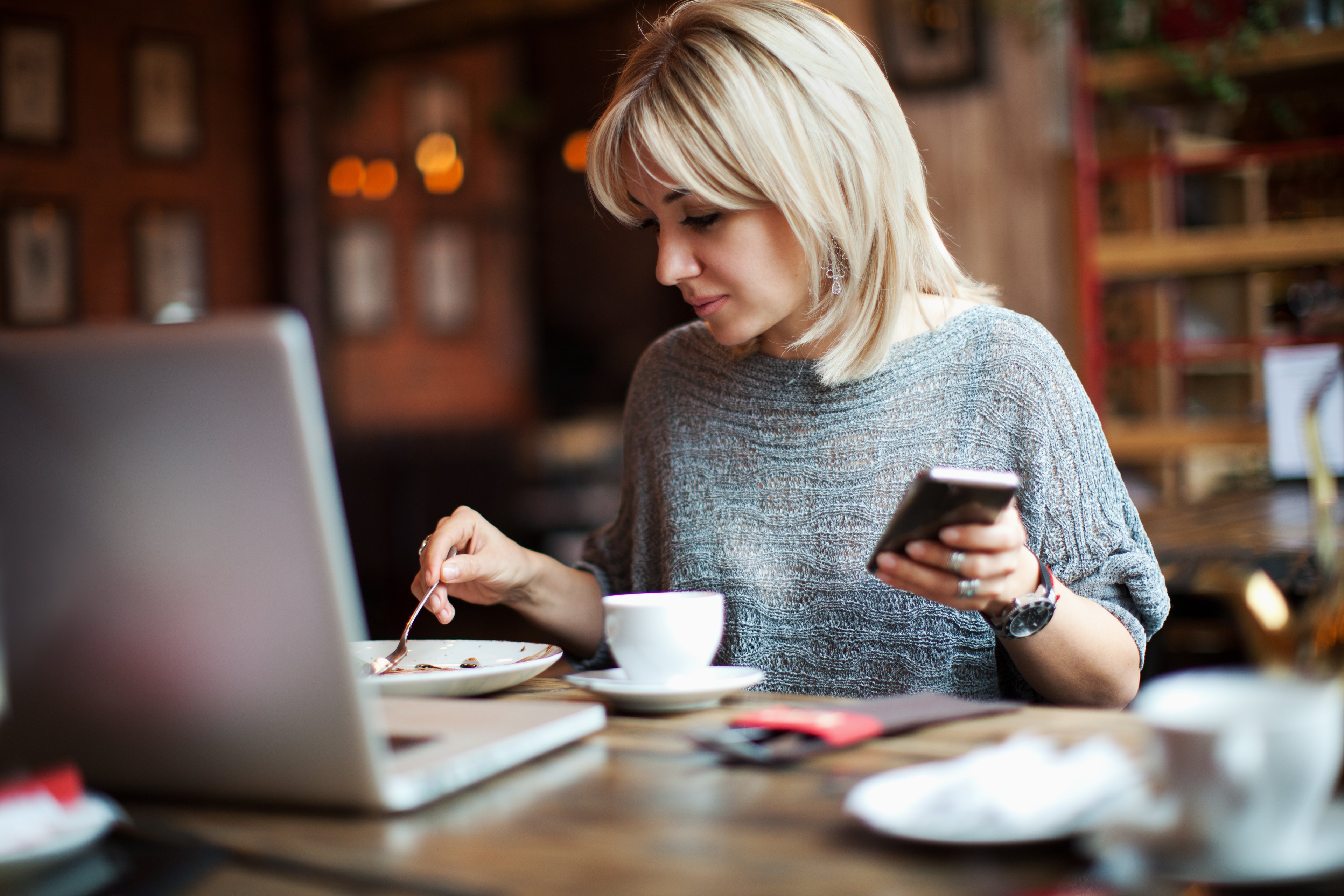 a woman sits at a table in a room holding a cell phone. On the table is a plate, a coffee cup and a laptop.